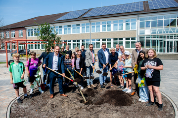 Wiebke Osigus steht in der Mitte einer Gruppe Erwachsener und Kinder, die Gruppe steht um ein Beet und schaut in die Kamera. Ein kleiner Baum liegt mit den Wurzeln in einem Erdloch. Die Erwachsenen halten Schaufeln mit Erde, um den Baum einzupflanzen.