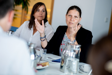 Ministerin Wiebke Osigus sitzt an einem Tisch und spricht mit einer Person, die ihr gegenüber sitzt. Neben ihr sitzt eine Frau und schaut sie an.