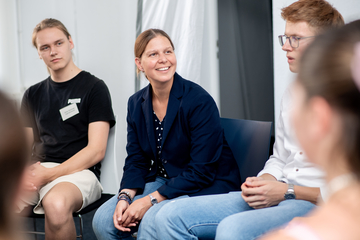 Ministerin Wiebke Osigus sitzt auf einem Stuhl, leicht vorgebeugt. Sie sitzt zwischen zwei männlichen Jugendlichen und lächelt. Sie schaut aus dem Bild heraus.