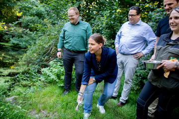 Die Ministerin Wiebke Osigus steht auf einer Wiese, die Knie leicht gebeugt, mit einem halben kleinen Fisch in der Hand. Um sie herum stehen vier Personen im Halbkreis.