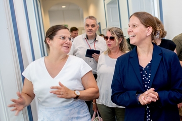 Eine Gruppe Menschen steht in einem hellen Flur mit hohen Decken. Die Ministerin steht vorne rechts im Bild und ist im Gespräch mit einer anderen Person. Sie lächelt und schaut auf etwas, das im Bild nicht zu sehen ist.