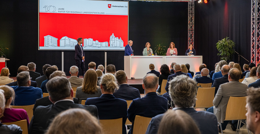 Im Hintergrund des Bildes ist eine Bühne mit großem Bildschirm zu sehen. Die vier Landesbeauftragten stehen an einem Podium, der Moderator steht etwas abseits. Im Vordergrund sitzt Publikum auf Stühlen, die Personen sind von hinten gezeigt.