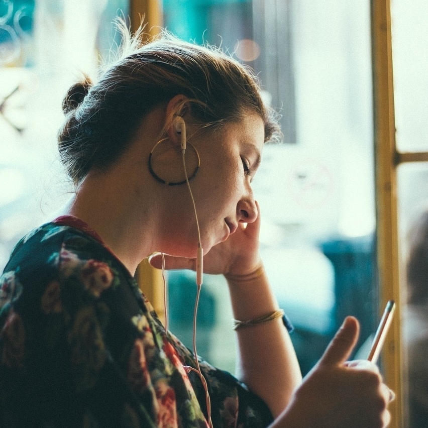 Eine junge Frau steht mit Kopfhörern im Ohr in einem öffentlichen Verkehrsmittel. Sie schaut mit ernstem Gesicht auf ihr Handy.