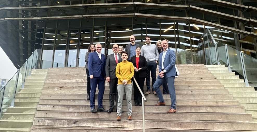 Die Ministerin Wiebke Osigus steht mit einer Gruppe Menschen im Freien auf einer Treppe. Im Hintergrund ist ein Gebäude mit großen Fenstern und einer Front aus Holzlamellen.