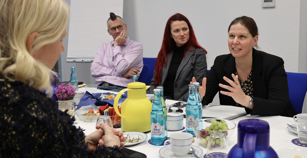 Eine Person (Ministerin Wiebke Osigus, rechts im Bild) im Gespräch mit einer weiteren Person (Barbara Gessler, vorne links im Bild, unscharf zu erkennen). Neben der Ministerin sitzen zwei Personen, die sie anschauen.