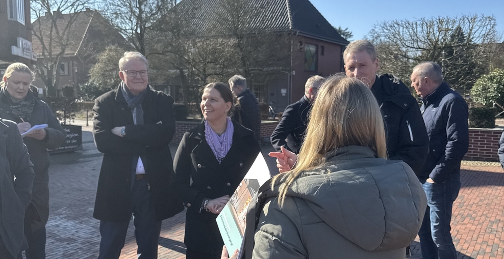 Ministerin Wiebke Osigus steht im Ortskern von Ganderkesee. Neben ihr stehen Nikolaus Jansen, Landesbeauftragter für regionale Entwicklung Weser-Ems, und Bürgermeister Ralf Wessel. Alle hören einer Mitarbeiterin der Gemeinde zu. Die Sonne scheint.